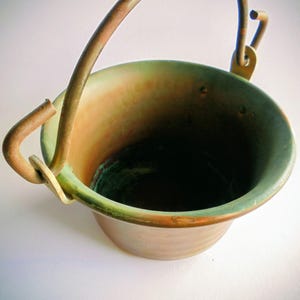 May include: A small, round, hammered metal bucket with a warm copper-toned exterior and a contrasting greenish-patina finish along the inner rim. It features a curved, arched metal handle attached to two side loops, creating a functional vessel.