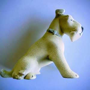 Pode incluir: Estatueta de c&atilde;o em cer&acirc;mica, em posi&ccedil;&atilde;o sentada, com acabamento texturizado em branco sujo. O c&atilde;o tem um nariz preto, uma coleira cinza e a boca ligeiramente aberta. A estatueta est&aacute; sobre um fundo azul claro.
