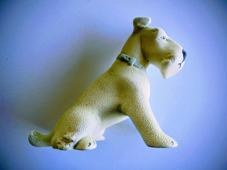 Pode incluir: Estatueta de c&atilde;o em cer&acirc;mica, em posi&ccedil;&atilde;o sentada, com acabamento texturizado em branco sujo. O c&atilde;o tem um nariz preto, uma coleira cinza e a boca ligeiramente aberta. A estatueta est&aacute; sobre um fundo azul claro.