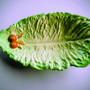 Op de afbeelding: Een keramieken serveerschaal in de vorm van een groot, gestructureerd groen slablad. De schaal heeft een glanzende afwerking met realistische gekreukte randen en een centrale nerf. Twee kleine, ronde, felrode tomaten zijn aan de zijkant bevestigd.
