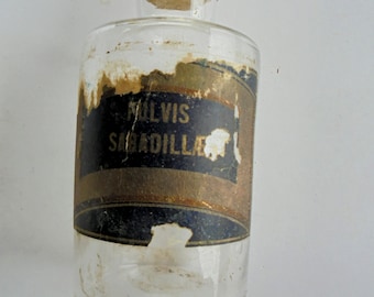 May include: A clear glass bottle with a cork stopper. The bottle has a label with the text "MILVIS SABADILLA" in black and gold. The label is partially torn and worn, giving it an antique appearance.