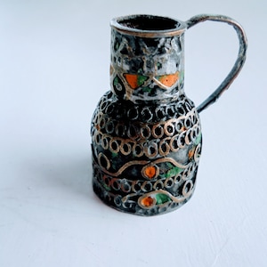 Può includere: Una piccola brocca antica color argento con manico. La brocca presenta un'intricata lavorazione traforata ed è adornata da accenti di smalto arancione e verde. Il design suggerisce un'origine vintage o artigianale, adatta all'esposizione o all'uso decorativo.