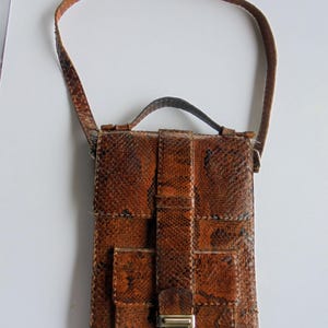 Puede incluir: Bolso bandolera vintage de cuero marrón con estampado de piel de serpiente. El bolso tiene forma rectangular con cierre de solapa, un asa pequeña y una correa larga. Tiene un cierre plateado.