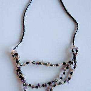 May include: A layered necklace featuring a dark gray beaded chain and three strands of amethyst and black gemstone beads. The necklace has small gold-colored spacer beads and is displayed on a white background.