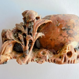 Könnte beinhalten: Eine geschnitzte Steinskulptur in Braun- und Beigetönen. Die Skulptur zeigt eine abgerundete Gefäßform mit strukturierter Oberfläche und ist mit floralen und Blattdetails verziert. Der Sockel hat einen geschnitzten, unregelmäßigen Rand.