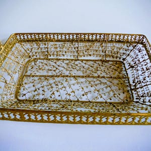 Op de afbeelding: Een rechthoekige mand van goudkleurig metaal met een ingewikkelde draadgaasbodem met een wervelend patroon. De mand heeft een decoratieve filigraanrand langs de bovenkant en twee gebogen handvatten aan de zijkanten, elk omwikkeld met spiraaldraad.