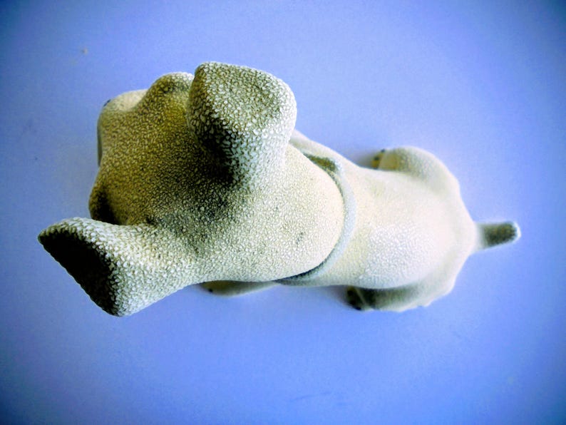 Pode incluir: Uma estatueta de c&atilde;o texturizada de cor clara, deitada de costas com as patas no ar. O c&atilde;o tem uma cauda pequena e uma coleira. O fundo &eacute; azul claro s&oacute;lido.