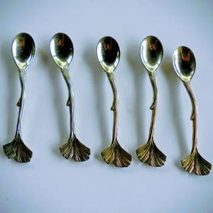 May include: A set of five silver-toned metal spoons arranged in a row. Each spoon features a smooth, oval-shaped bowl and a handle designed to resemble a slender stem that terminates in a textured, fan-shaped ginkgo leaf at the base.