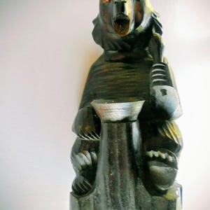 Peut inclure: Figurine d'ours en bois sculpté avec des yeux rouges et une bouche ouverte. L'ours tient un récipient et est peint dans des tons de vert, de marron et de noir. La figurine est posée sur un petit socle.