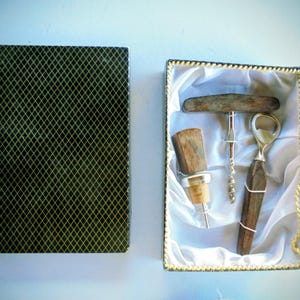 Puede incluir: Un juego de accesorios para vino en una caja rectangular con tapa de patrón de diamantes negro y dorado. El interior, forrado en satén blanco, contiene un tapón de madera, un sacacorchos en T y un abridor con mango de madera oscura.