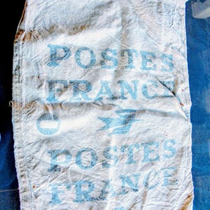 May include: A vintage, off-white canvas mail sack with the words "POSTES FRANCE" printed in faded blue. The sack has a drawstring closure and shows signs of wear, suggesting age and use. The French postal service logo is also visible.
