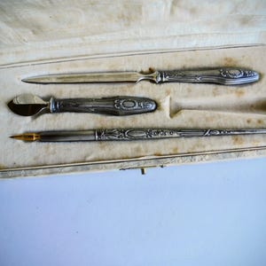 Op de afbeelding: Een set van drie zilverkleurige bureauaccessoires, waaronder een briefopener, een lakstempel en een vulpen met een goudkleurige penpunt. Elk item heeft een gedetailleerd, reliëf handvat en zit in een crèmekleurige, met fluweel beklede doos.