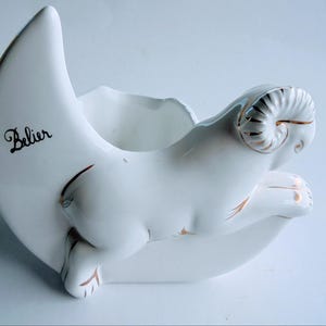 May include: White ceramic Aries zodiac planter in the shape of a crescent moon. The ram figure has gold accents on its horns and hooves. The word "Belier" is written in black script on the moon.