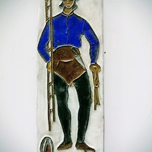May include: A rectangular ceramic tile featuring a raised relief illustration of a figure in a blue jacket, brown apron, and black leggings. The figure holds a tall ladder and a rope. The tile includes a small emblem and the text NAMUR 84.