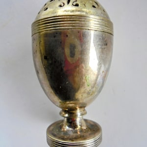 May include: An antique silver-coloured spice shaker, egg-shaped with a perforated top. It has a pedestal base and horizontal ridged bands around the top and base. The metal shows signs of age and wear, with a tarnished appearance.