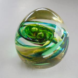 Könnte beinhalten: Ein eiförmiger Glas-Briefbeschwerer mit wirbelnden grünen, blauen und gelben Mustern. Das klare Glas hat eine glänzende Oberfläche, die das Licht reflektiert und die farbenfrohen Muster hervorhebt. Ein dekorativer Artikel.