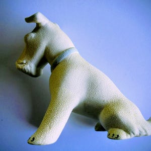Pode incluir: Estatueta de c&atilde;o em cer&acirc;mica creme com superf&iacute;cie texturizada. O c&atilde;o est&aacute; sentado com a cabe&ccedil;a levantada e tem uma coleira azul. A estatueta tem detalhes pretos nas patas, sobre um fundo azul.