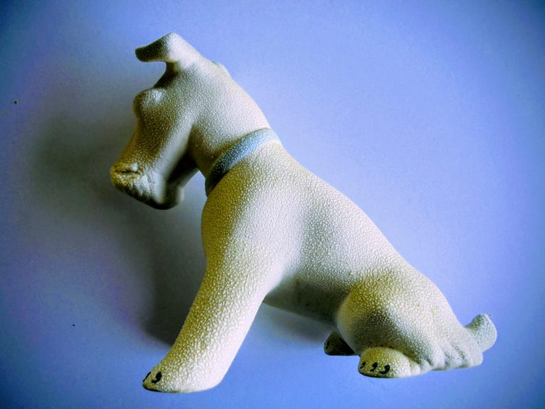 Pode incluir: Estatueta de c&atilde;o em cer&acirc;mica creme com superf&iacute;cie texturizada. O c&atilde;o est&aacute; sentado com a cabe&ccedil;a levantada e tem uma coleira azul. A estatueta tem detalhes pretos nas patas, sobre um fundo azul.