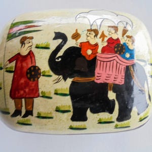May include: Hand-painted rectangular box with a cream-colored background. The box features a black elephant carrying people with a red-clad figure on the left. The figures are dressed in bright colors, and the scene is reminiscent of an Indian procession.