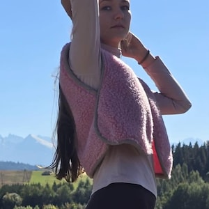 Può includere: Un gilet senza maniche rosa chiaro realizzato in un materiale felpato testurizzato e soffice. Il gilet presenta un design semplice aperto sul davanti con un bordo grigio a contrasto lungo i profili. È indossato sopra un top beige a collo alto.