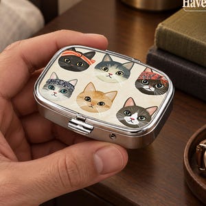 Op de afbeelding: Een rechthoekig metalen pillendoosje met een zilveren afwerking, voorzien van een geprint patroon van zes gestileerde kattengezichten op het deksel. De katten zijn afgebeeld in diverse kleuren, waaronder zwart, oranje en grijs, met hoofdbanden.