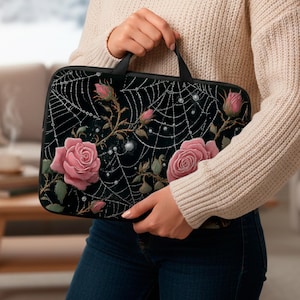 Op de afbeelding: Zwarte laptoptas met een ontwerp van roze rozen, spinnenwebben en dauwdruppels. De rozen staan in volle bloei en de spinnenwebben zijn gedetailleerd met kleine, glinsterende kralen. De tas heeft een zwart handvat.