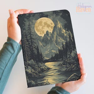 Op de afbeelding: Een notitieboek met een landschapsillustratie van een volle maan boven bergen, een rivier en een bos. Het kunstwerk gebruikt grijstinten, zwart en geel. Het notitieboek wordt door een persoon vastgehouden.