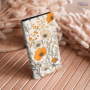 Peut inclure: Une coque de téléphone rectangulaire avec un fond crème orné d'un motif répétitif de fleurs sauvages orange et gris sourd, incluant des coquelicots et des tiges délicates. Le design recouvre la surface avant d'une illustration botanique.