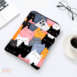 May include: A rectangular laptop sleeve featuring a colorful pattern of smiling cartoon cats in various colors, including black, pink, orange, and gray. A white smartphone is partially visible inside the sleeve. The sleeve is on a white surface.