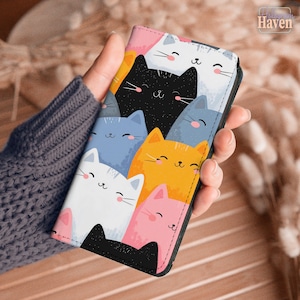 May include: A rectangular phone case featuring a colorful pattern of cartoon cat faces in white, black, blue, orange, and pink. Each cat has simple line-drawn eyes, a small nose, and pink cheeks. The design covers the entire front surface.