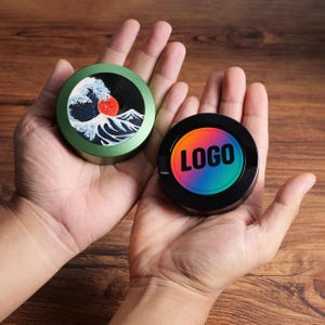May include: Two circular containers are held in open hands. One is green with a wave and red sun design. The other is black with a rainbow gradient and the word "LOGO".