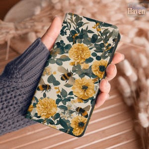 Puede incluir: Una funda rectangular para teléfono con fondo color crema y un patrón repetitivo de flores amarillas, hojas verde oscuro y abejorros. El diseño incluye varias abejas en vuelo entre los elementos florales, creando un tema botánico.