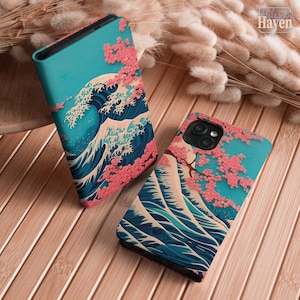 May include: Two rectangular phone cases featuring a graphic print of a large dark blue ocean wave with white foam crests against a bright turquoise sky, accented by clusters of pink cherry blossoms on branches extending across the surface.