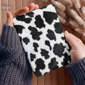 May include: A rectangular item with a cowhide pattern, featuring black spots on a white background. The item is held by hands wearing a gray sweater. The item has a soft, textured appearance.