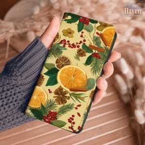 May include: A phone case featuring a festive design with orange slices, pine needles, red berries, and cinnamon sticks on a cream background. The case has a black border and a decorative pattern, perfect for the holiday season.