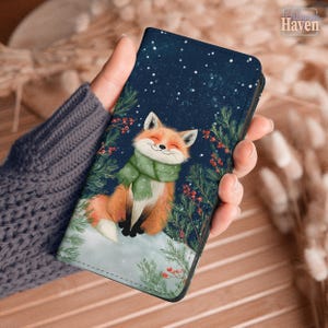 May include: A phone case featuring a smiling fox wearing a green scarf, set against a snowy winter scene with a starry night sky. The fox is orange and white, surrounded by green pine branches and red berries.