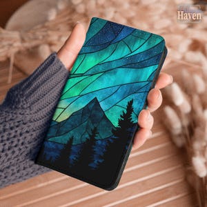 May include: A rectangular phone case featuring a stained glass style design of a mountain range and pine trees under a vibrant teal and blue aurora borealis sky filled with tiny white stars. The artwork uses dark lines to create geometric shapes.