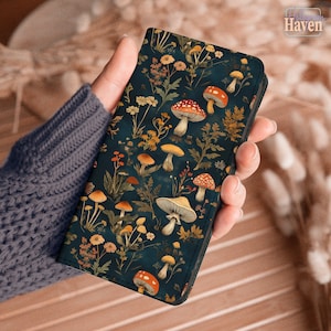 May include: A dark teal wallet featuring a whimsical mushroom and floral pattern. The design includes various mushroom illustrations in shades of red, orange, and yellow, set against a backdrop of foliage and flowers. The wallet is held in a person's hand.