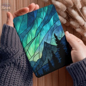 May include: A tablet case featuring a stained-glass design of the Northern Lights. The artwork depicts a vibrant aurora borealis in shades of teal and green, with a starry sky and silhouetted mountains and trees.