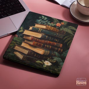 Può includere: Una custodia decorativa per laptop con una pila di libri antichi con pagine color oro e copertine in pelle scura. Il design include fogliame verde e piccoli fiori, creando un'estetica vintage. La custodia è su una superficie rosa.