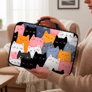 Könnte beinhalten: Eine schwarze Laptoptasche mit einem sich wiederholenden Muster aus lächelnden Cartoon-Katzen in verschiedenen Farben, darunter Rosa, Orange, Weiß, Schwarz und Blau. Die Tasche hat einen schwarzen Griff und einen schwarzen Rand.