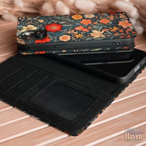 May include: A floral patterned phone case with a wallet design. The case has a dark blue background with red, orange, and yellow flowers. The wallet section is black and includes card slots and a clear ID window. The phone is black.