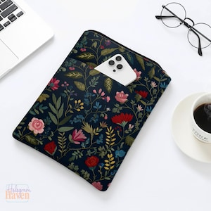 May include: A navy blue laptop sleeve with a floral design of pink, red, and blue flowers and green leaves. A white smartphone is in the front pocket. The sleeve is on a white surface.