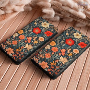 May include: Two phone cases with a floral design. The cases have a dark teal background with red, orange, and yellow flowers and green leaves. They are displayed on a wooden surface.