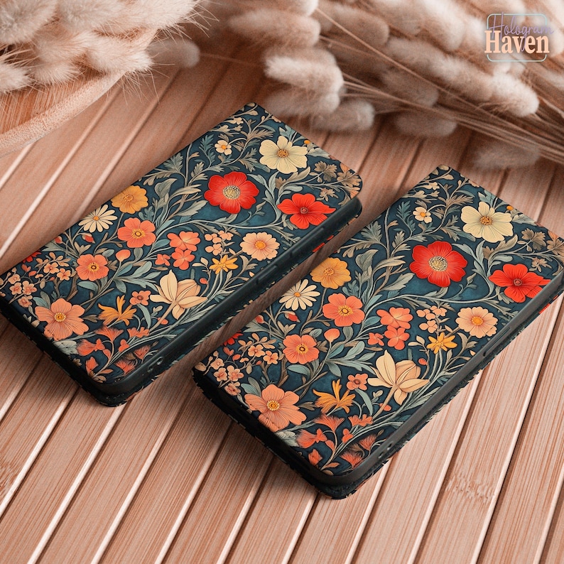 May include: Two phone cases with a floral design. The cases have a dark teal background with red, orange, and yellow flowers and green leaves. They are displayed on a wooden surface.