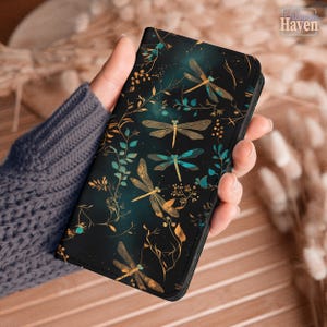 Puede incluir: Una funda de teléfono negra con un diseño de libélulas. La funda tiene un fondo oscuro con libélulas doradas y turquesas, hojas turquesas y enredaderas doradas. La funda está en la mano de una persona.