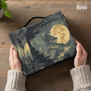 May include: A black and gray landscape-themed handbag featuring a full moon, mountains, and trees. The bag has a black handle and the text "Hologram Haven" in the upper right corner.