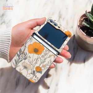 May include: A floral patterned phone case for a Samsung Galaxy Z Flip. The case features a cream background with orange poppies and other flowers. The phone case is open, revealing the phone's screen and the Samsung logo.