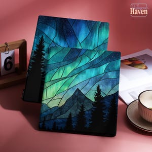 May include: Two tablet cases with a stained glass design depicting an aurora borealis scene. The design features shades of blue, green, and black, with a mountain range and silhouetted trees. The cases are displayed on a pink surface.