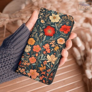May include: A phone case with a dark teal floral design, featuring red, orange, yellow, and white flowers. The case is held in a hand, highlighting the detailed pattern and colour scheme. The case has a black border.